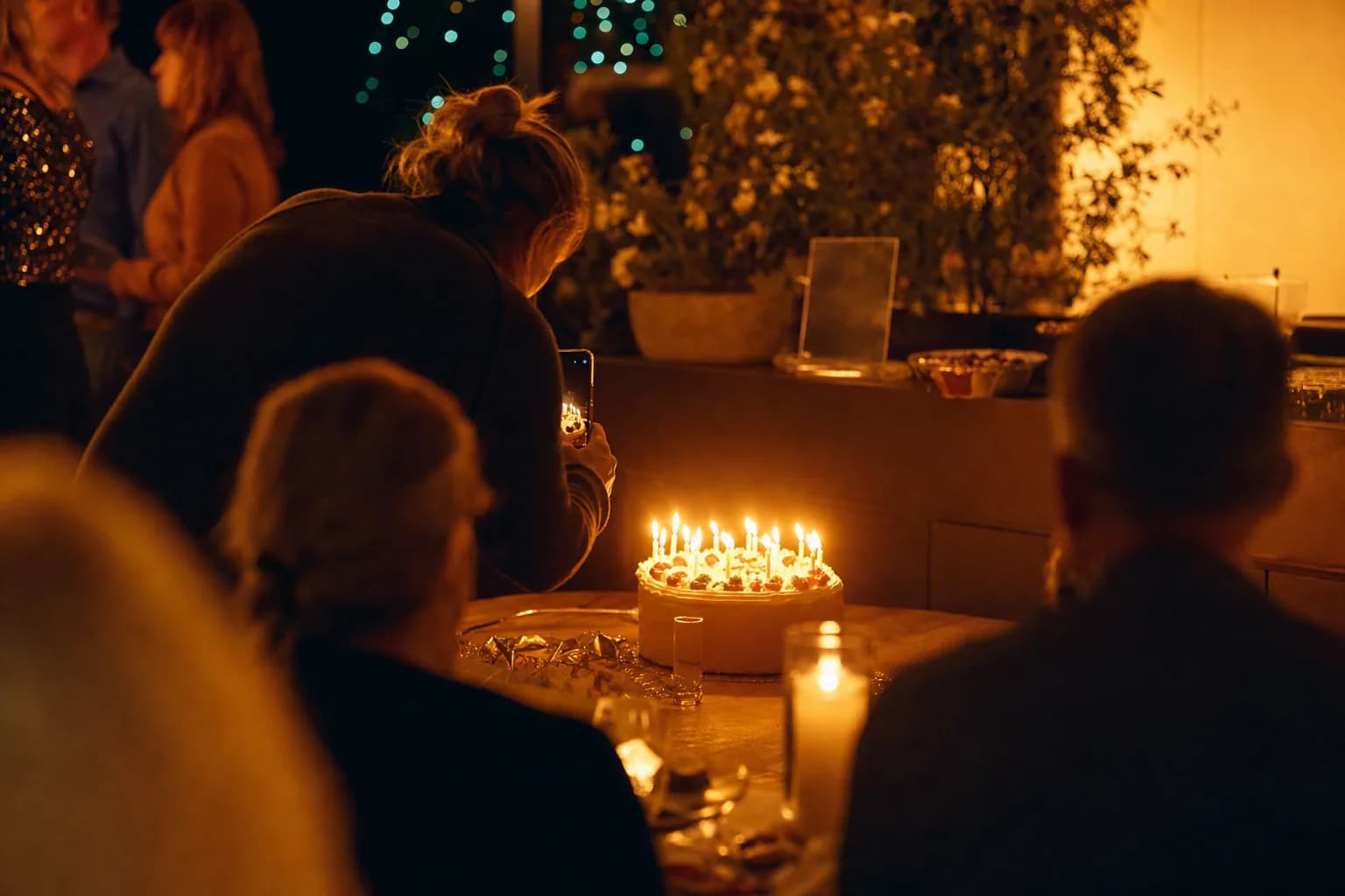 Geburtstagsfeier im Warmlicht: jemand fotografiert eine Torte mit brennenden Kerzen, feiernde Gäste unscharf im Hintergrund, warmes Kerzenlicht vor dunklem Grund.