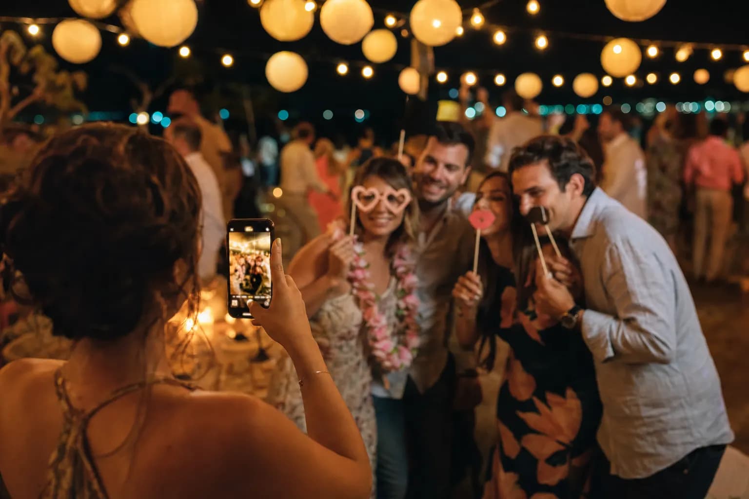 Freunde posieren auf einer Feier ausgelassen mit Papier-Props für ein Smartphone-Foto — der Fotobox-Moment ganz ohne Fotobox, mitten im warmen Partylicht. (KI-generiertes Bild)