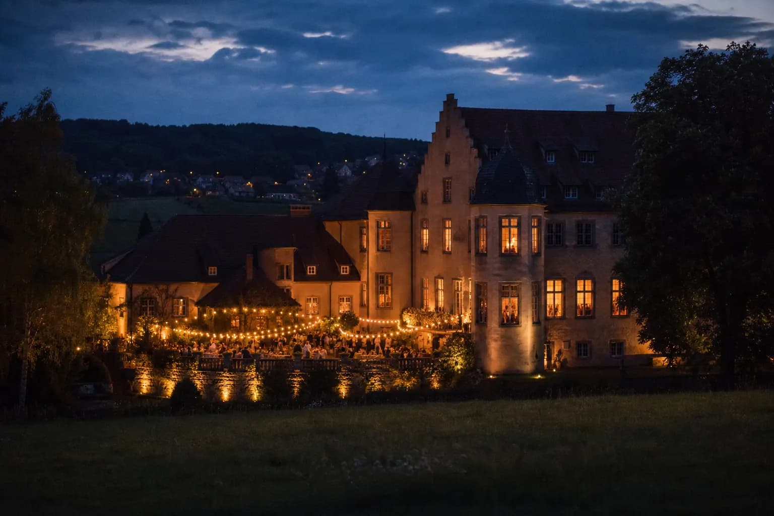 Ein warm leuchtendes Feier-Anwesen in der deutschen Abendlandschaft, auf Terrasse und hinter den Fenstern feiern Gäste als kleine warme Silhouetten. (KI-generiertes Bild)