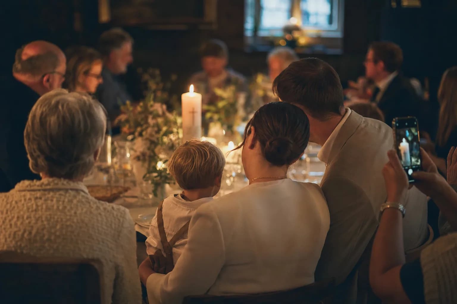 Behutsame Tauffeier: eine Taufkerze leuchtet warm, drei Generationen einer Familie von hinten an der Festtafel, sanftes goldenes Kirchenlicht vor ruhigem, dunklem Grund.
