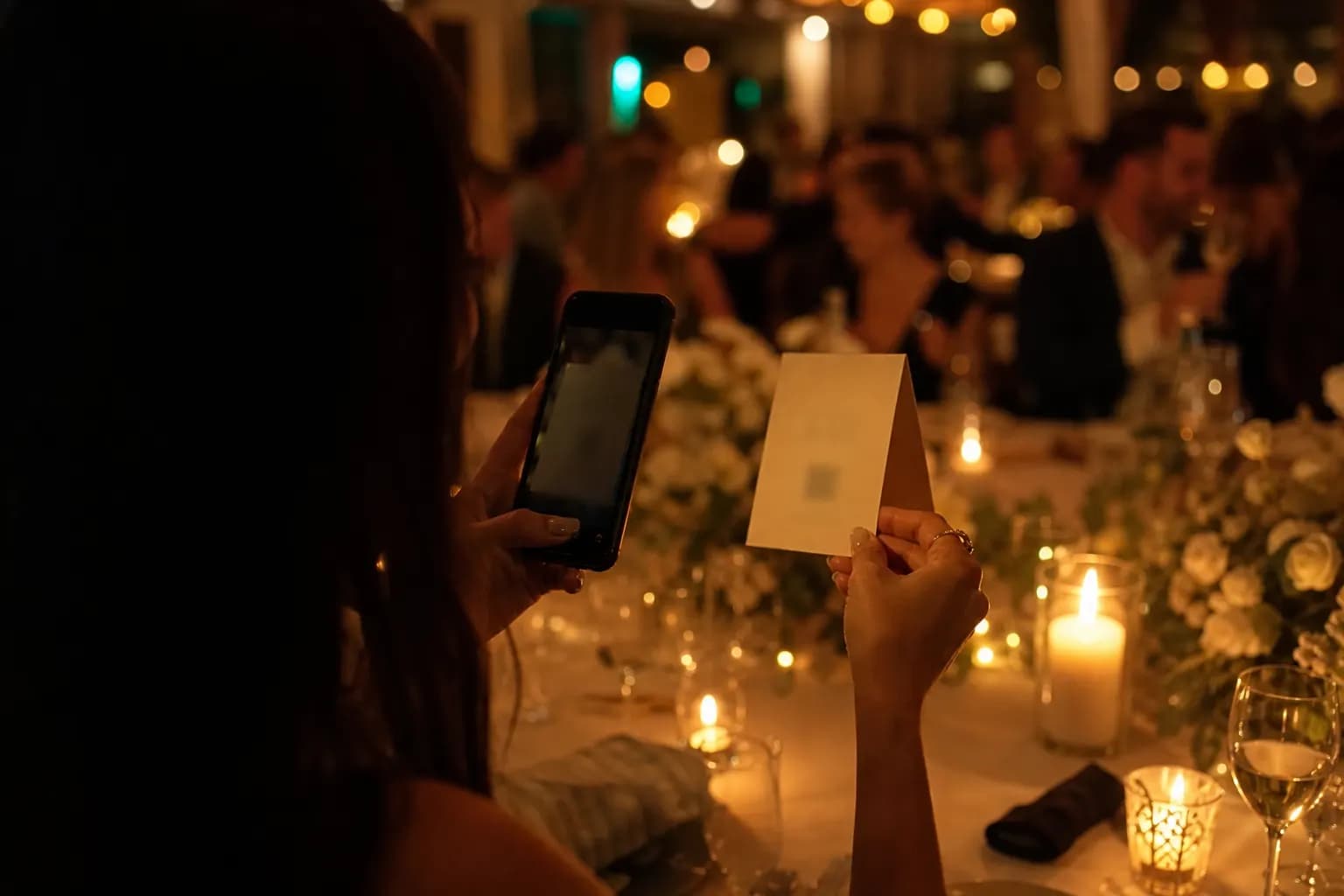 Ein Gast hebt auf einer warm beleuchteten Feier eine kleine Tischkarte an und richtet sein Smartphone darauf, im Hintergrund feiern Gäste.